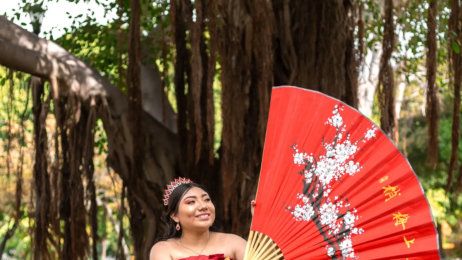 Quinceañera con vestido rojo y corona sentada en el pasto junto a un abanico rojo gigante pintado con flores, bajo árboles