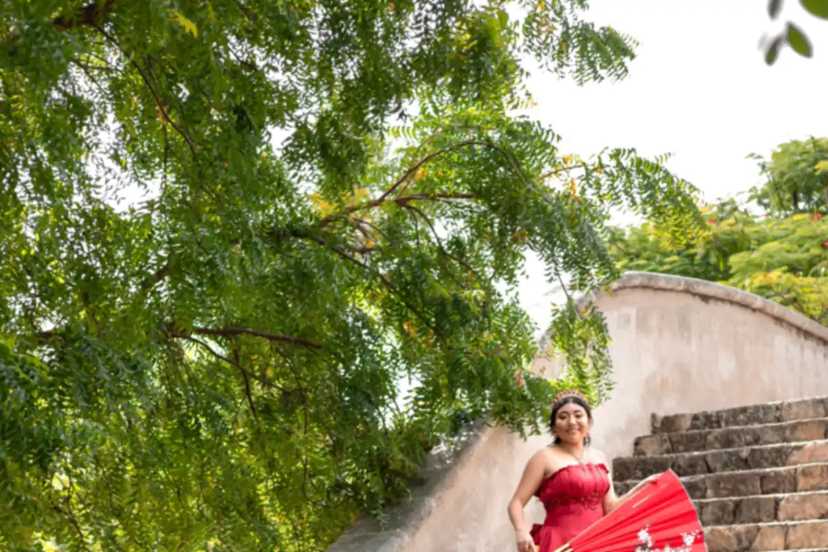 Quinceañera con vestido rojo en unas escaleras de piedra rodeadas de árboles, con un abanico rojo
