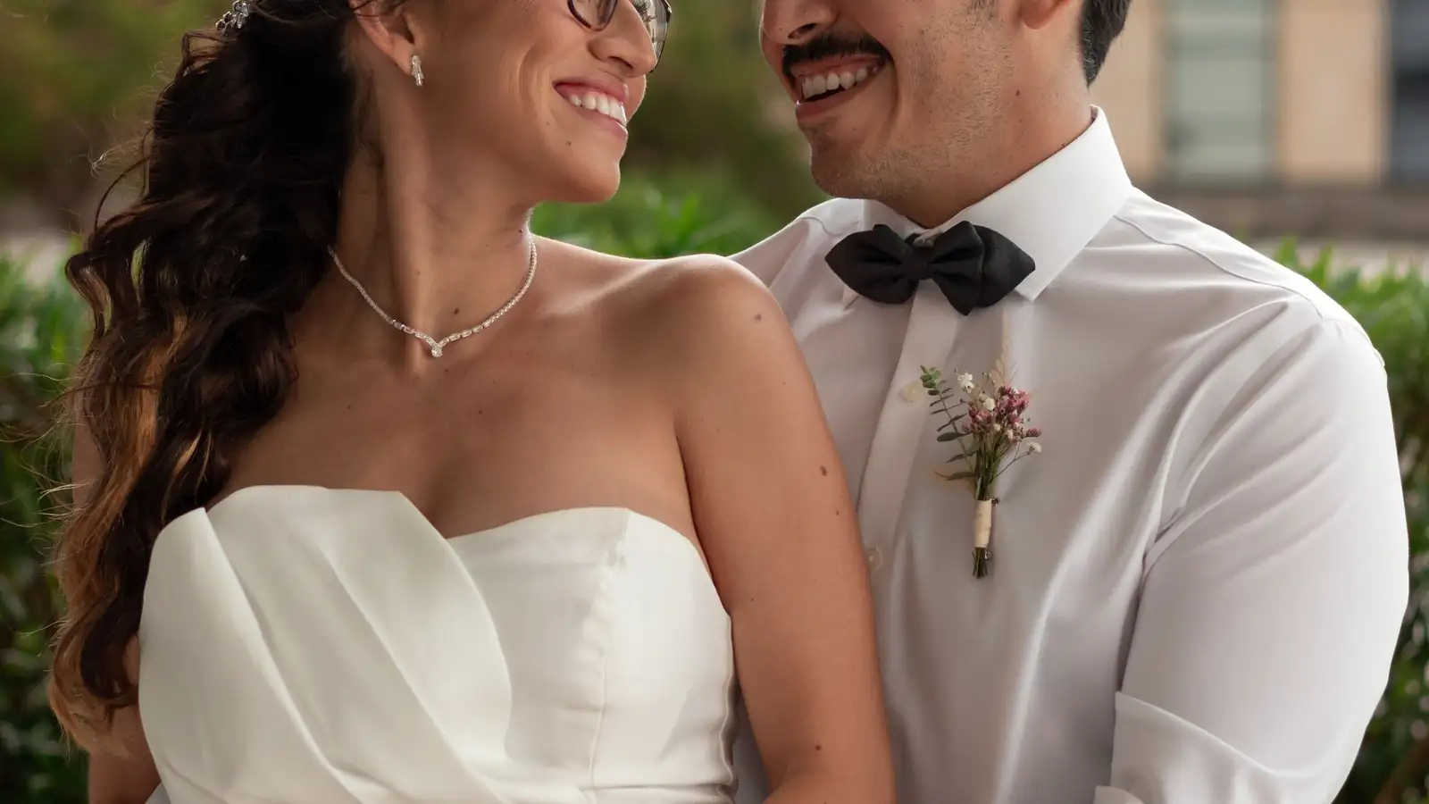 Novios sonriendo muy cerca uno del otro, él con moño negro y ella con vestido strapless