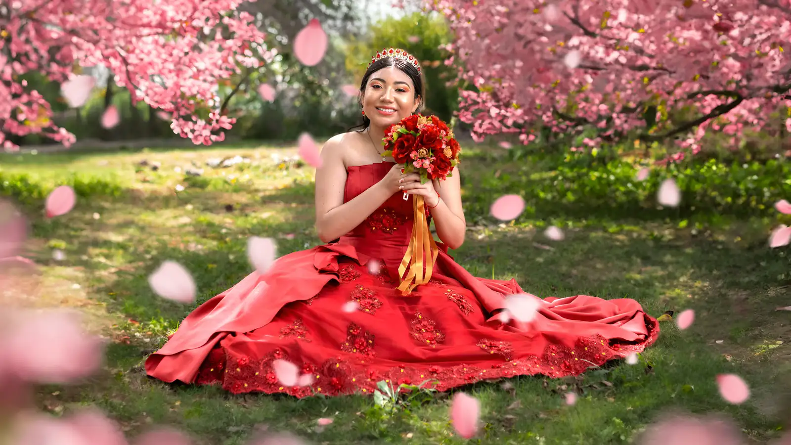 Quinceañera con vestido rojo sentada en el pasto bajo un árbol de flores rosas, con su ramo en las manos