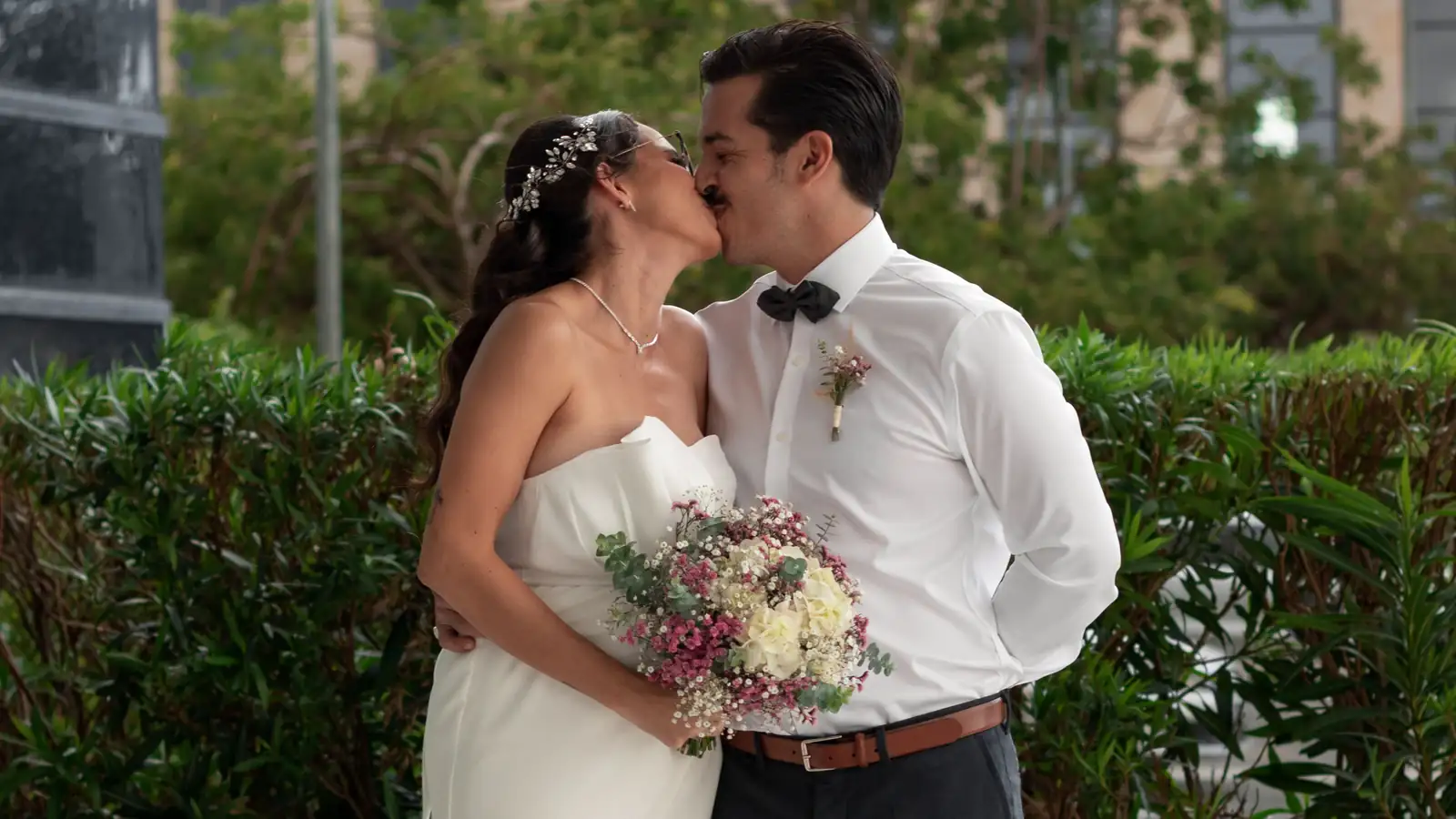Novios besándose en un jardín después de su boda civil, ella con vestido blanco sencillo y ramo de flores