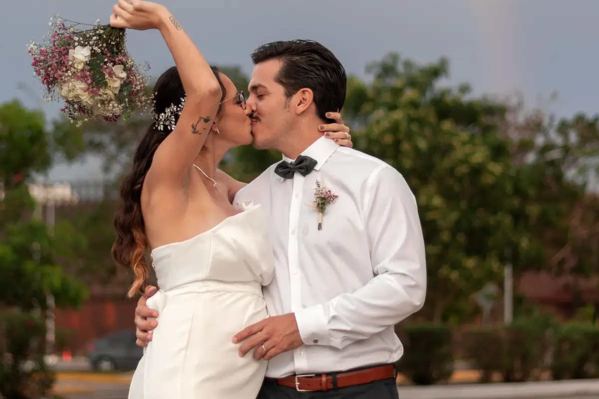 Novios besándose al aire libre con un arcoíris de fondo, ella levantando su ramo