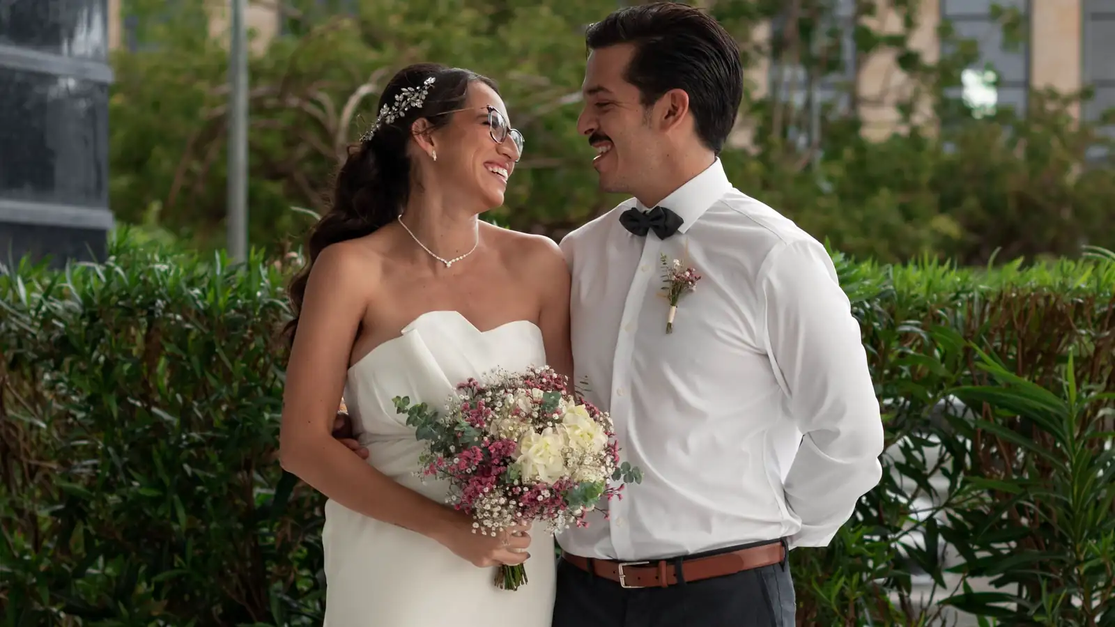 Novios abrazados en un jardín después de su boda civil, ella con vestido sencillo y un ramo de flores