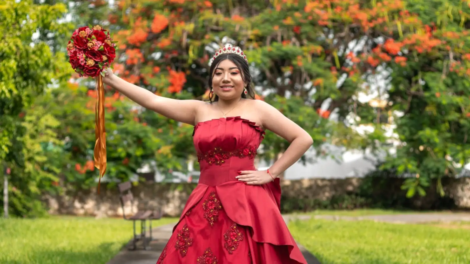 Quinceañera de vestido rojo en un camino de jardín, con el ramo en alto y un árbol de flores rojas detrás