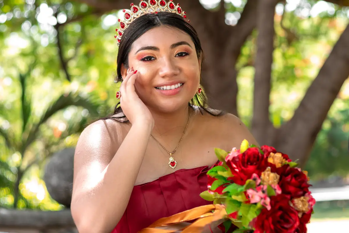 Quinceañera sentada en el pasto bajo los árboles, junto a un gran abanico rojo con flores