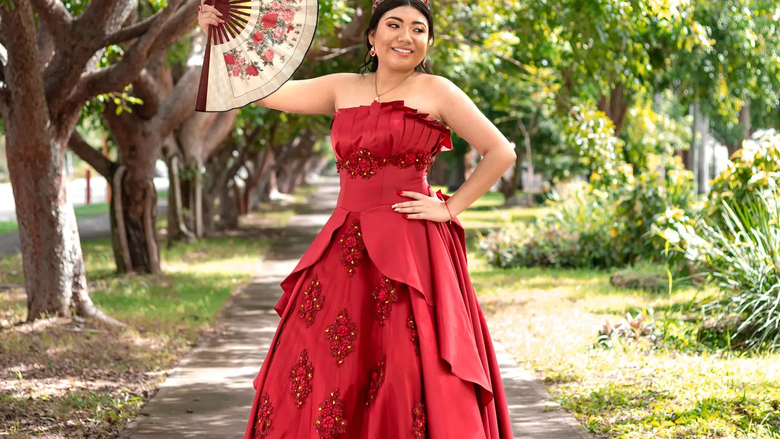 Quinceañera con vestido rojo y corona caminando por un sendero entre árboles con un abanico en la mano