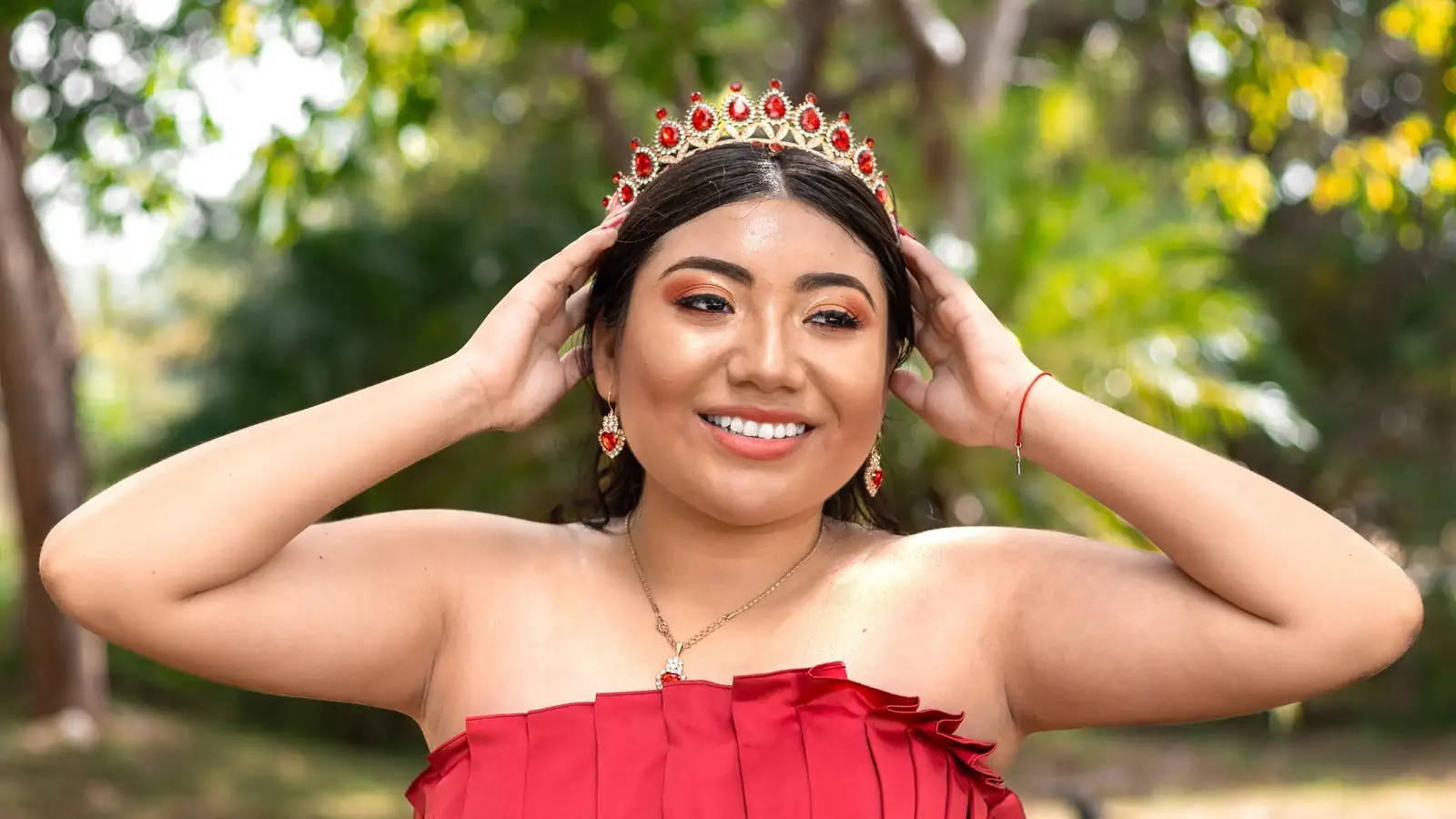 Quinceañera de vestido rojo sonriendo con las manos sobre su corona