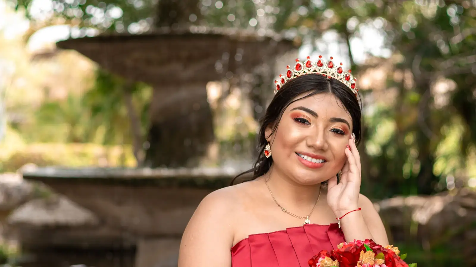 Quinceañera sentada junto a una fuente con su ramo de rosas rojas y corona