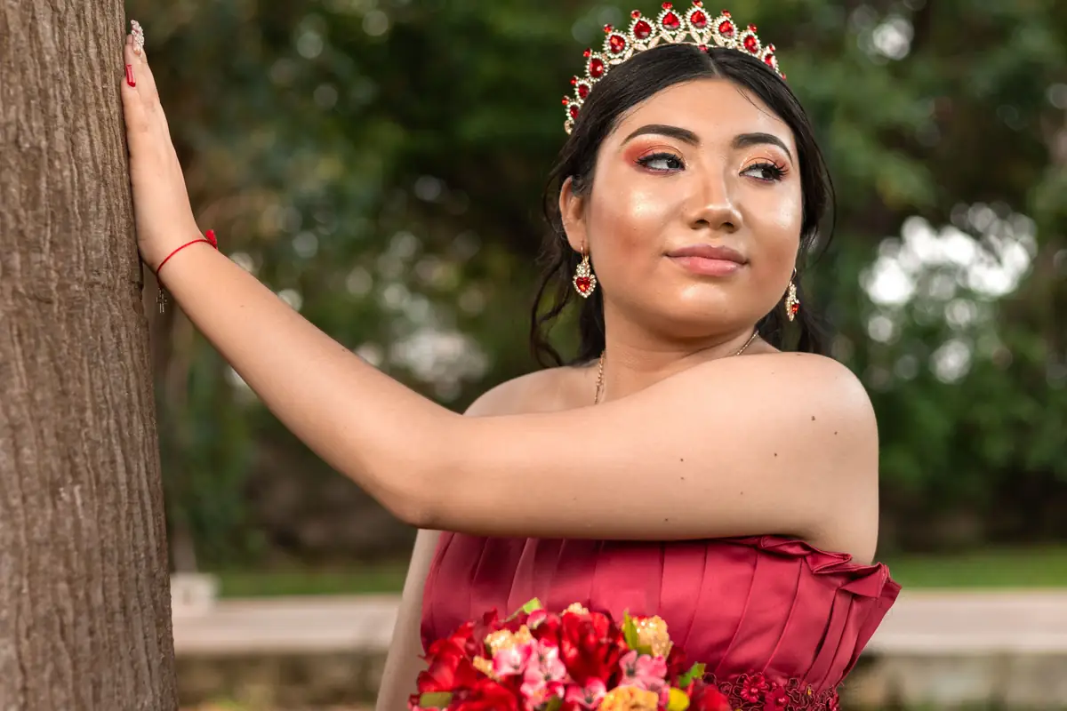 Quinceañera recargada en un árbol con su ramo de rosas rojas, sonriendo