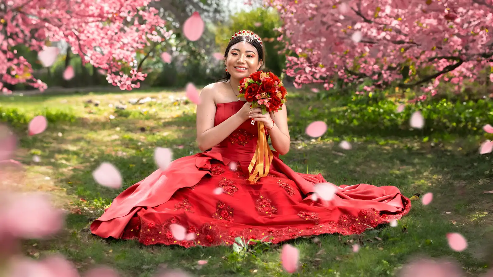 Quinceañera con vestido rojo y ramo de flores sentada en el pasto entre pétalos rosas