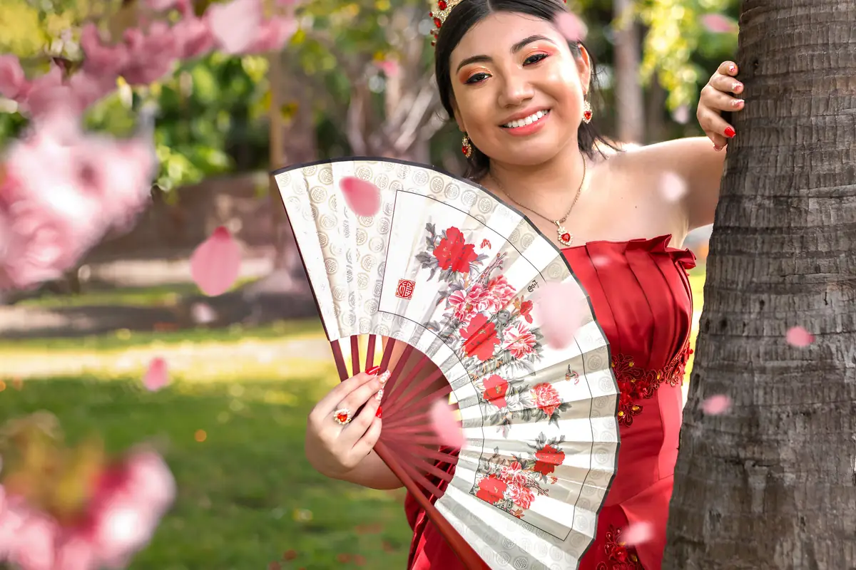 Quinceañera sonriente con vestido rojo y un abanico de flores, recargada en un árbol