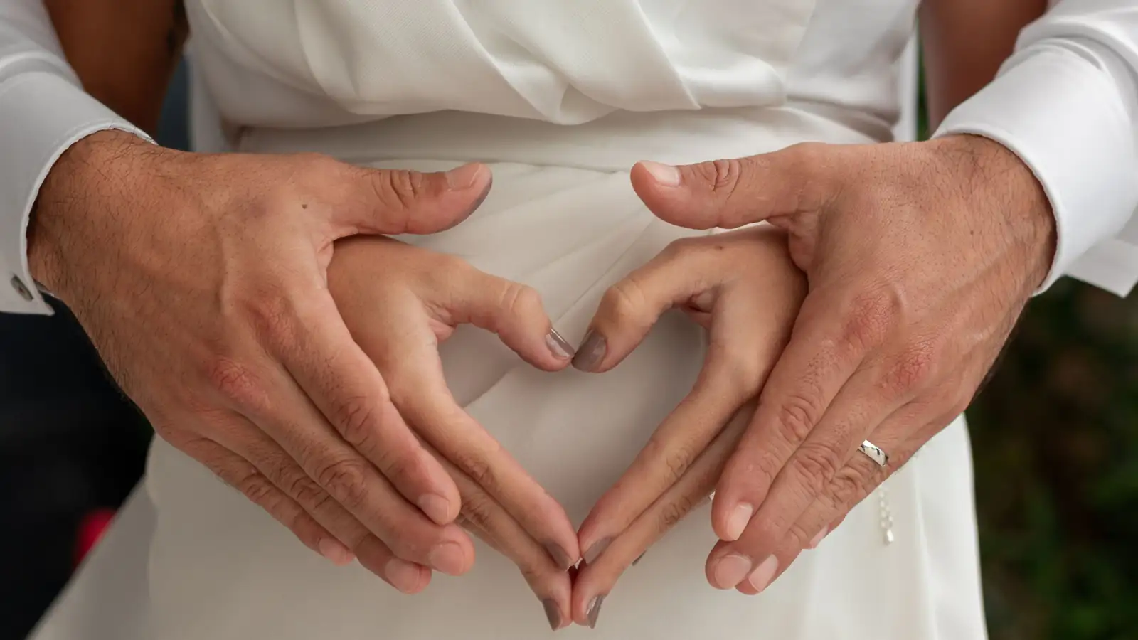 Manos de los novios formando un corazón sobre el vestido blanco, con sus anillos de boda a la vista